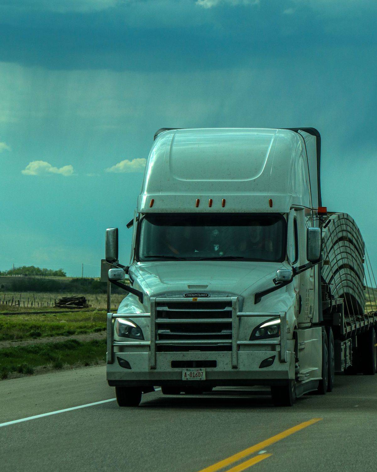 White Freightliner tractor hauling a flatbed load on a two-lane highway under a storm sky